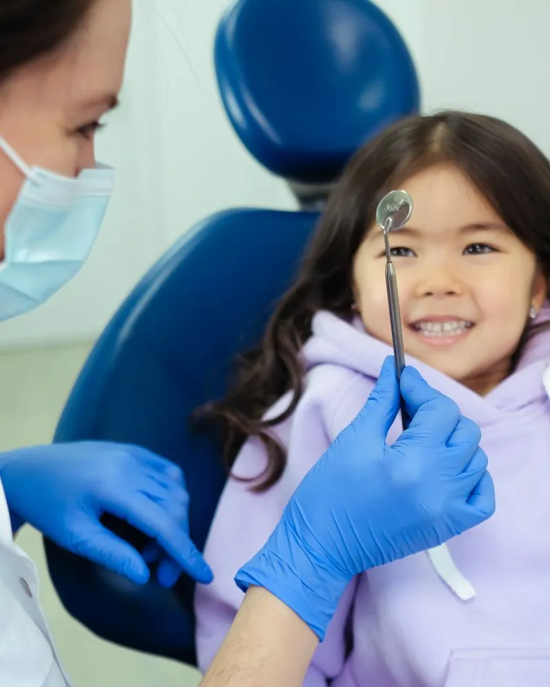 A young girl smiles happily in a dental chair as her dentist holds a mirror during a routine visit.